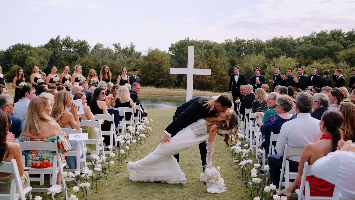 Wedding guests seated on white chairs watch a couple share a special moment in front of a cross at an outdoor ceremony.