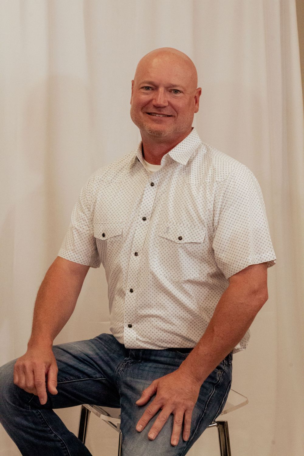 A person in a white button-down short sleeve shirt and jeans sits casually on a chair against a light backdrop.