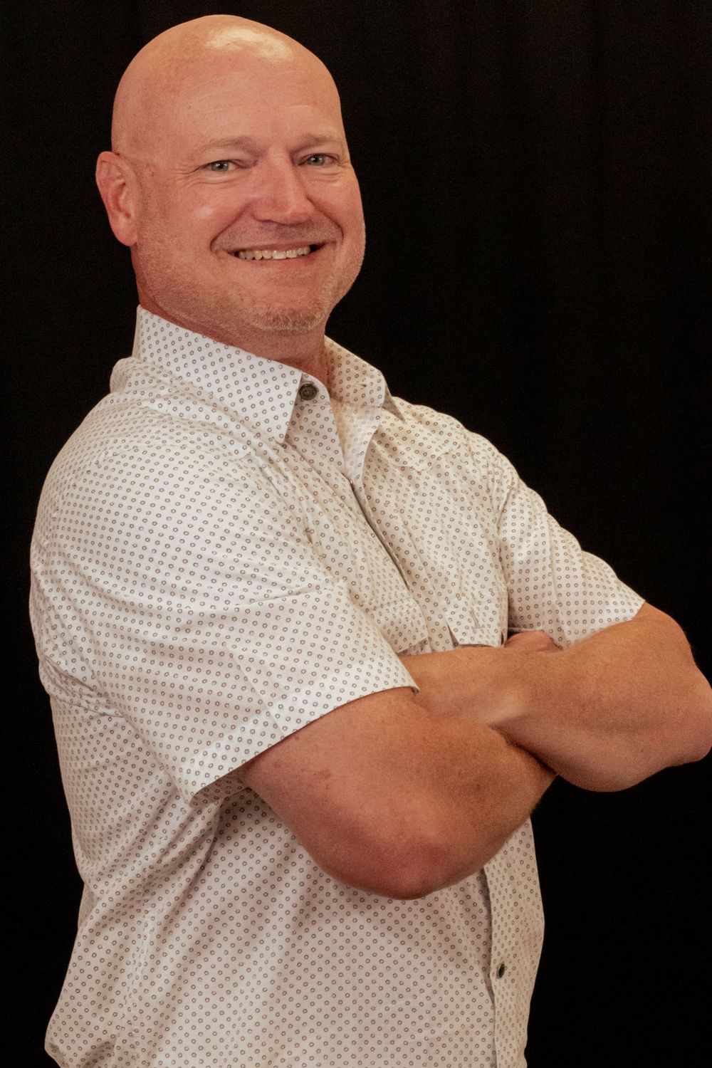 Professional headshot of a bald person in a white polka dot dress shirt with arms crossed against a black background.