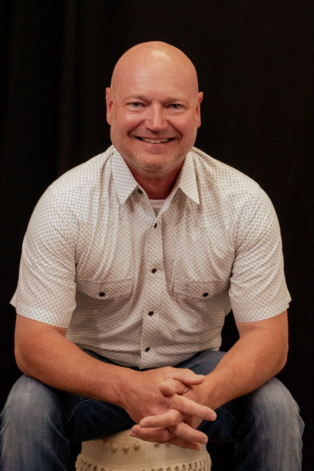A bald man in a white patterned short-sleeve shirt and jeans sits casually while smiling at the camera.