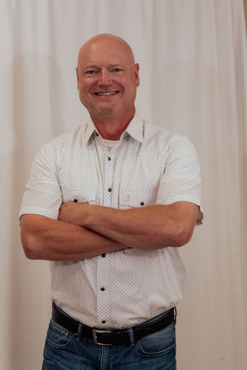 A person in a white patterned button-down shirt and jeans stands confidently with arms crossed against a light backdrop.