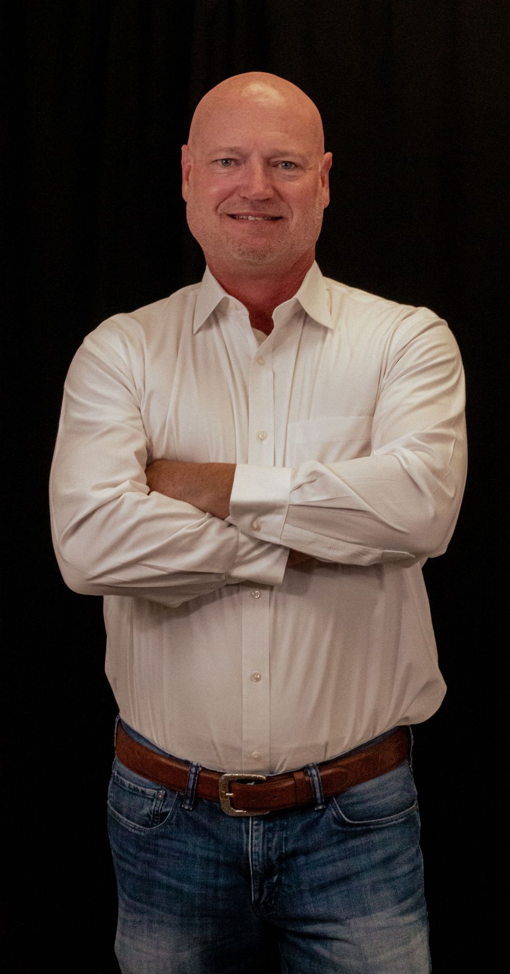 Person wearing a white button-down shirt and blue jeans poses with arms crossed against dark background.