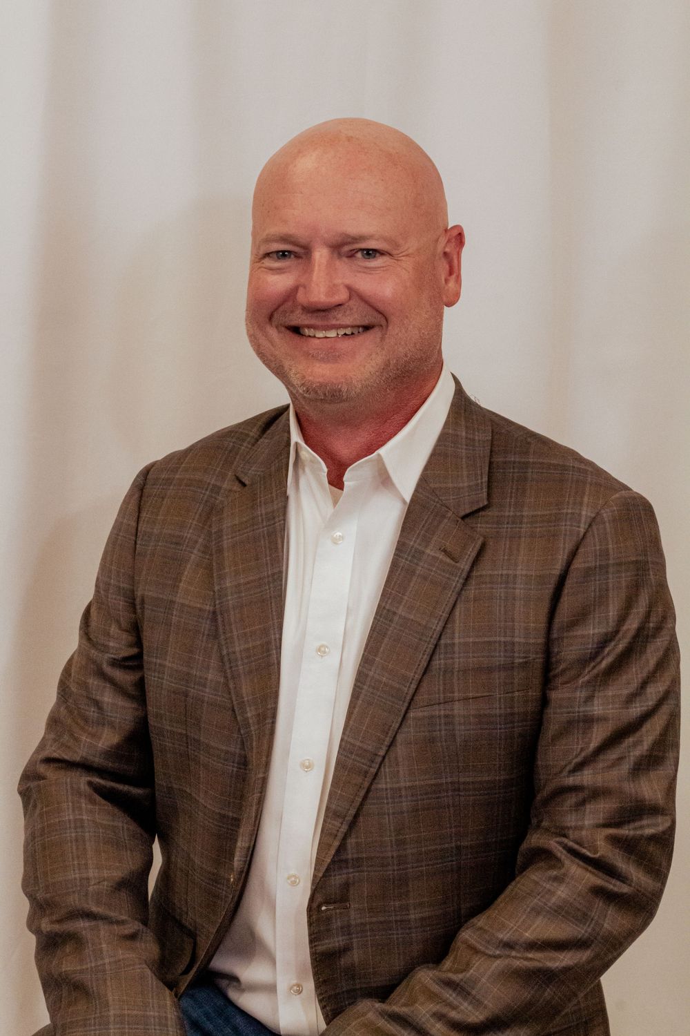 Professional business headshot of man in brown plaid blazer and white dress shirt against light background.