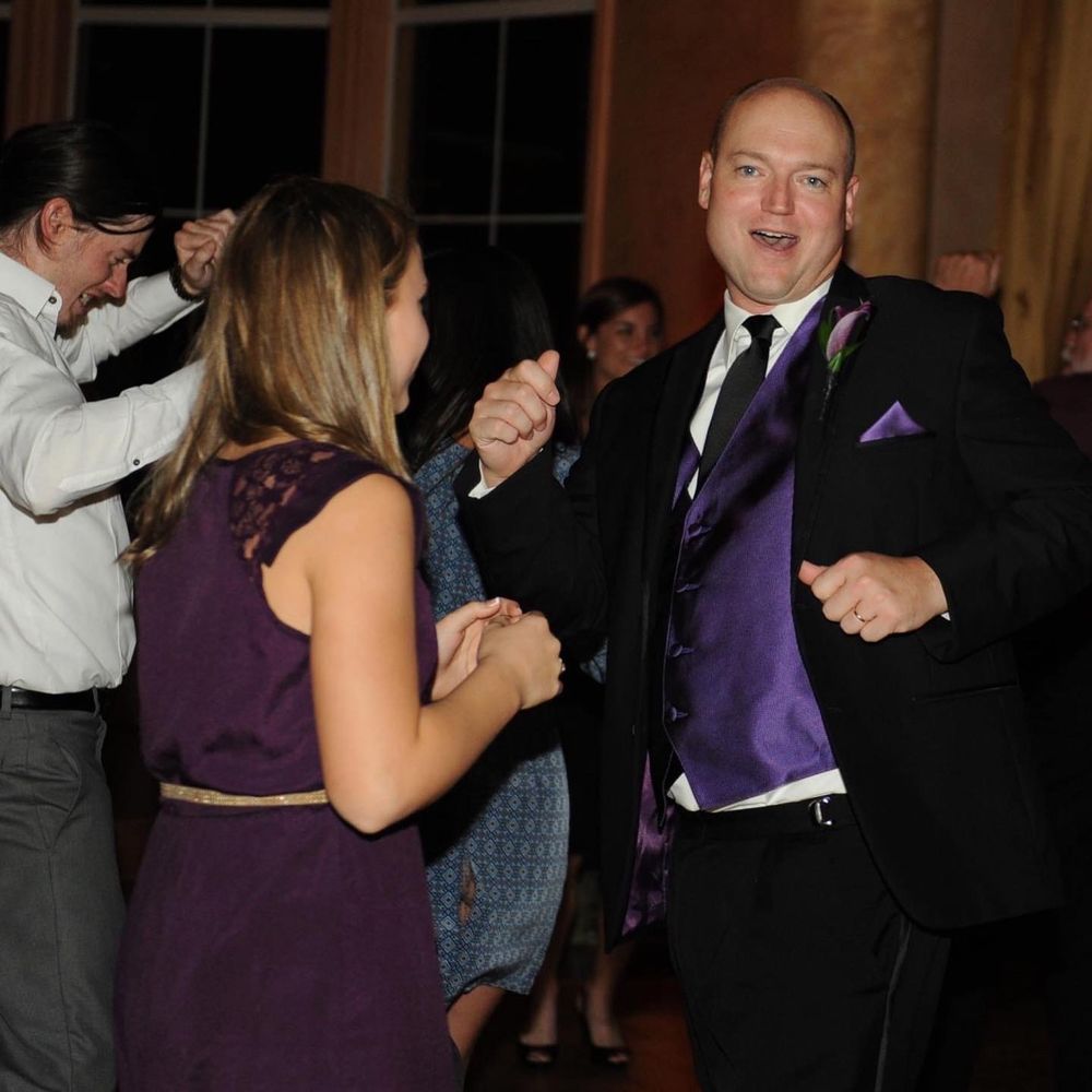 A couple dancing together at an elegant event, with the man wearing a black suit and purple accessories.