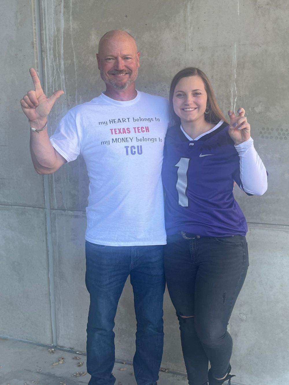 Two people in casual attire standing against a concrete wall making peace signs while smiling at the camera.