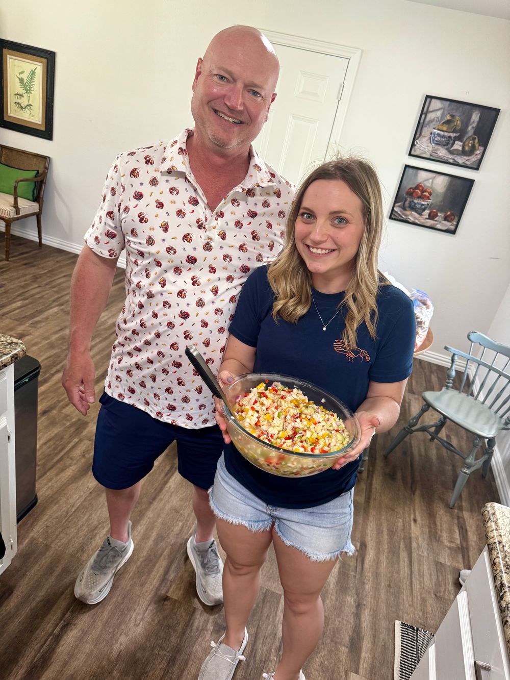 Two people posing with a colorful dessert dish in a home interior with wood floors and framed artwork on the walls.