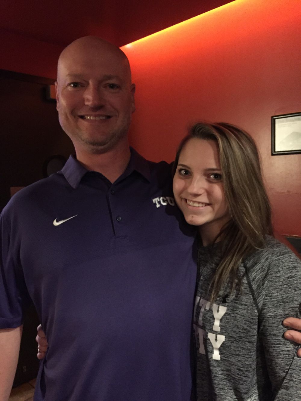 Two people wearing casual athletic wear pose together indoors against a wall with warm red lighting.