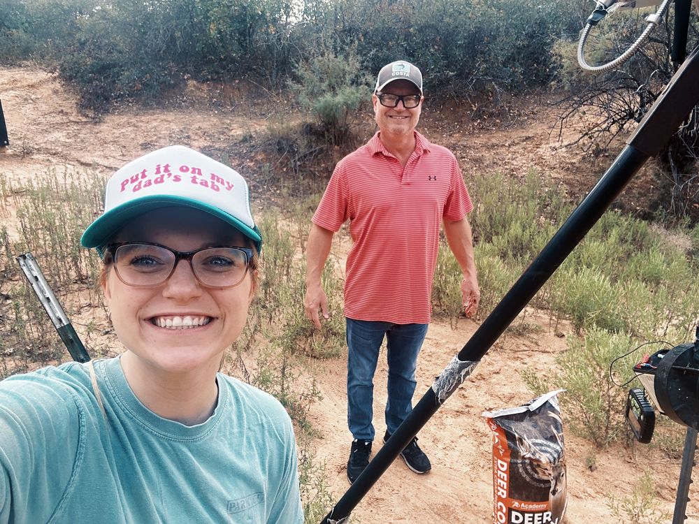 A hiking selfie taken on a dirt trail with two people wearing casual outdoor attire and baseball caps.