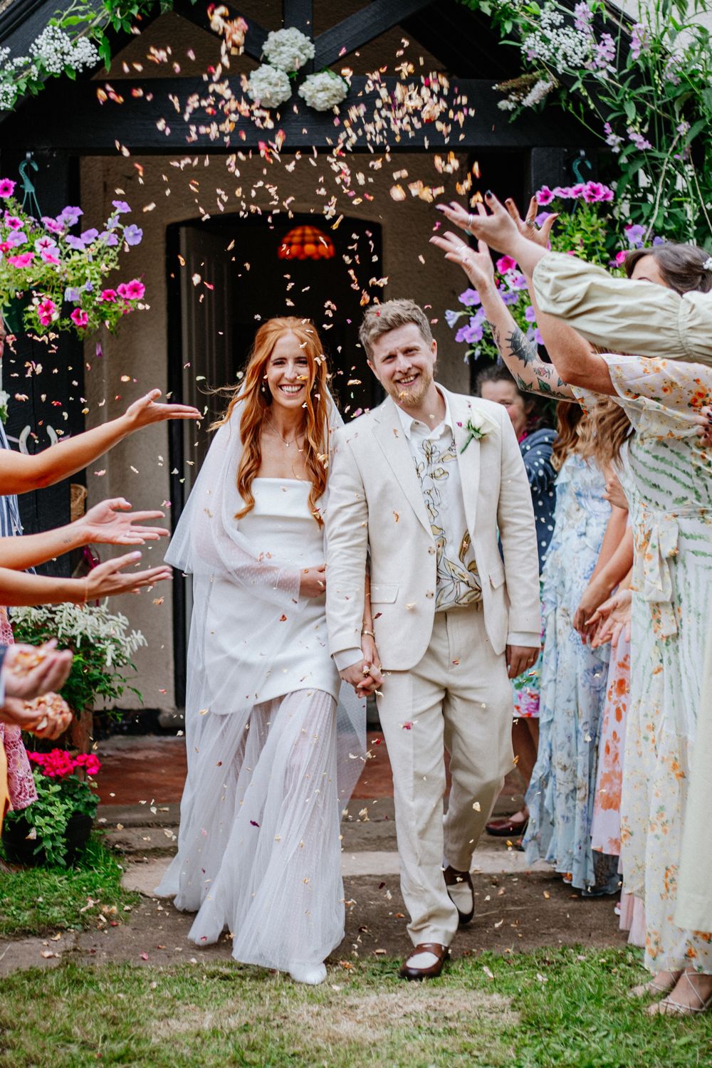 Newlyweds walking down garden path while guests shower them with confetti at outdoor wedding celebration.