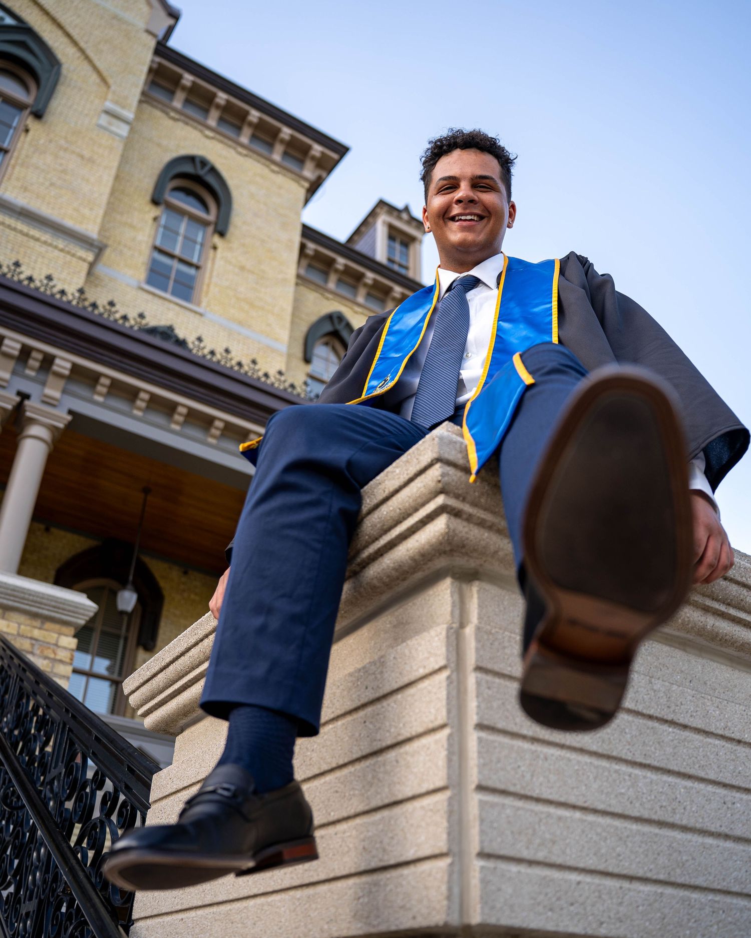 Graduate wearing a black cap and gown with blue and gold stoles poses under a stone archway during commencement.