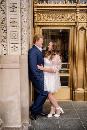 A couple in wedding attire poses playfully in front of an ornate stone building with gold-trimmed doors.