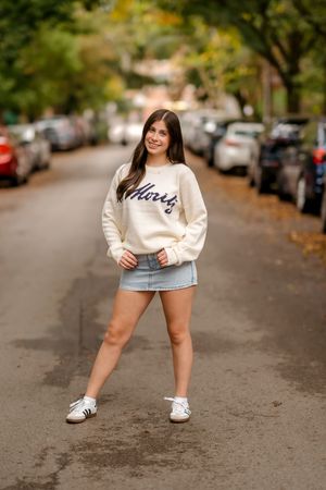 Young woman in cream sweatshirt and denim mini skirt poses on a tree-lined street with parked cars in autumn.