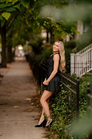 Smiling blonde woman in black mini dress posing on a tree-lined sidewalk next to an iron fence.