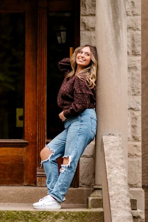 Young woman in ripped jeans and dark floral top leaning against stone building, smiling upward.