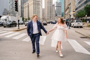 A couple in wedding attire dances playfully across a city crosswalk with tall buildings in the background.