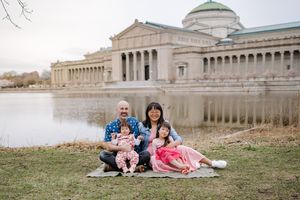 Family of four poses by a pond with a neoclassical museum building in the background on a cloudy day.
