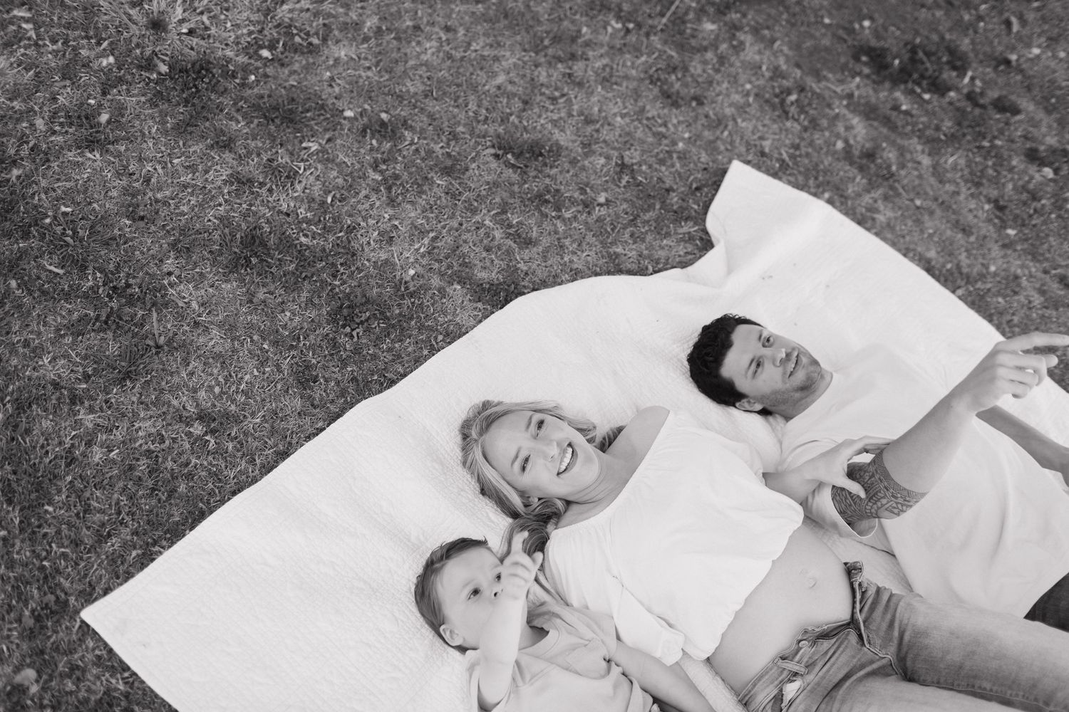 Three people laying together on a white blanket on the grass, shot from above in black and white.