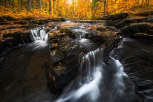 Golden autumn sunlight illuminates a cascading waterfall flowing over dark rocky terrain in a forest.