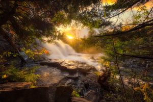 Golden sunlight breaks through forest mist above dramatic waterfall at sunset.
