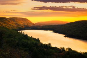 Panoramic sunset view over a tranquil lake surrounded by rolling mountains with orange and purple sky.