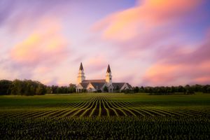 White church with twin spires stands against dramatic sunset sky over neat rows of crops in a rural field.