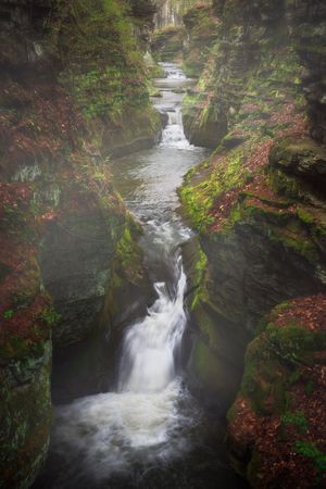 A dramatic waterfall flows through a narrow rocky gorge surrounded by autumn foliage and moss-covered cliffs.