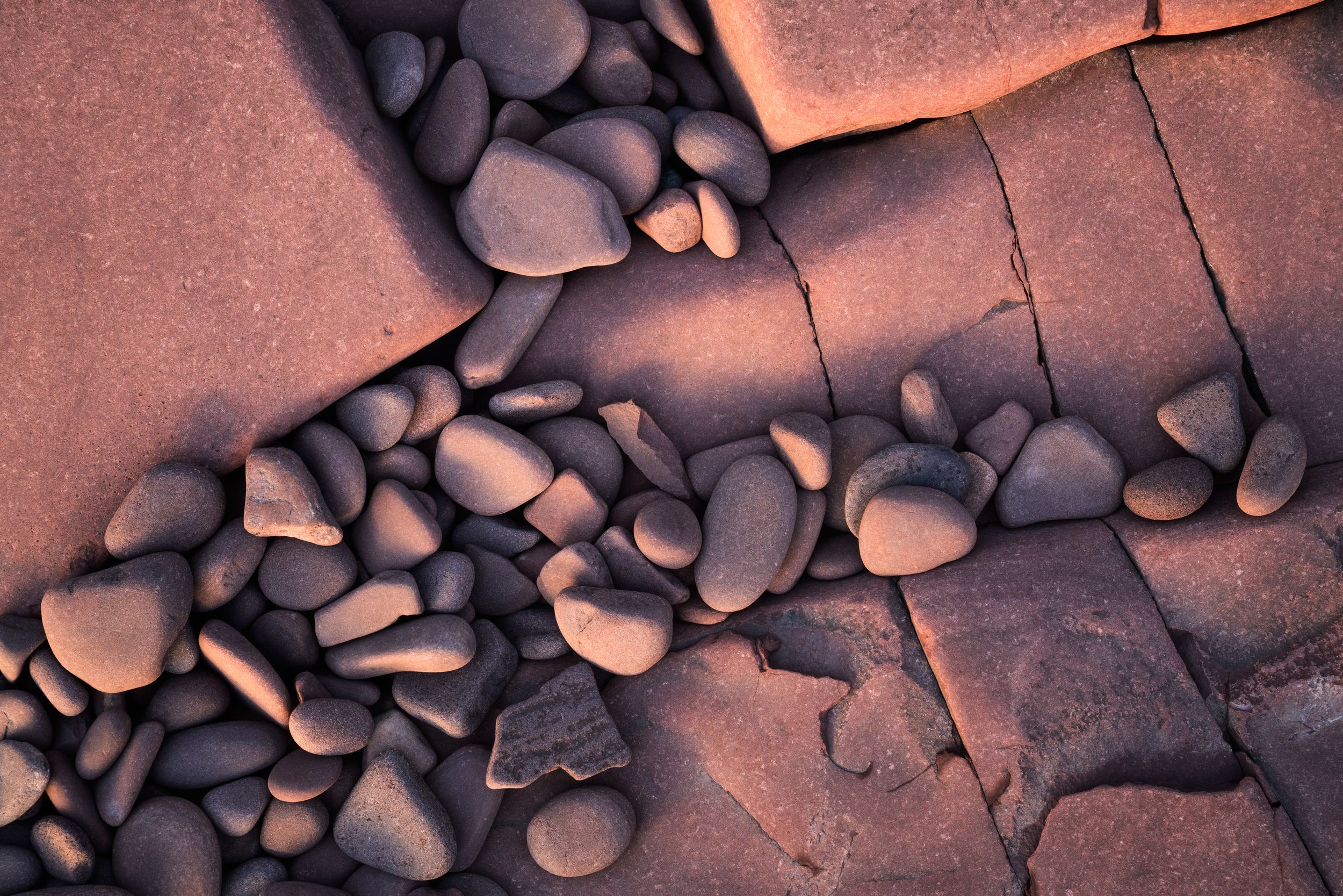 Smooth river rocks scattered across cracked red sandstone create an abstract natural pattern at sunset.