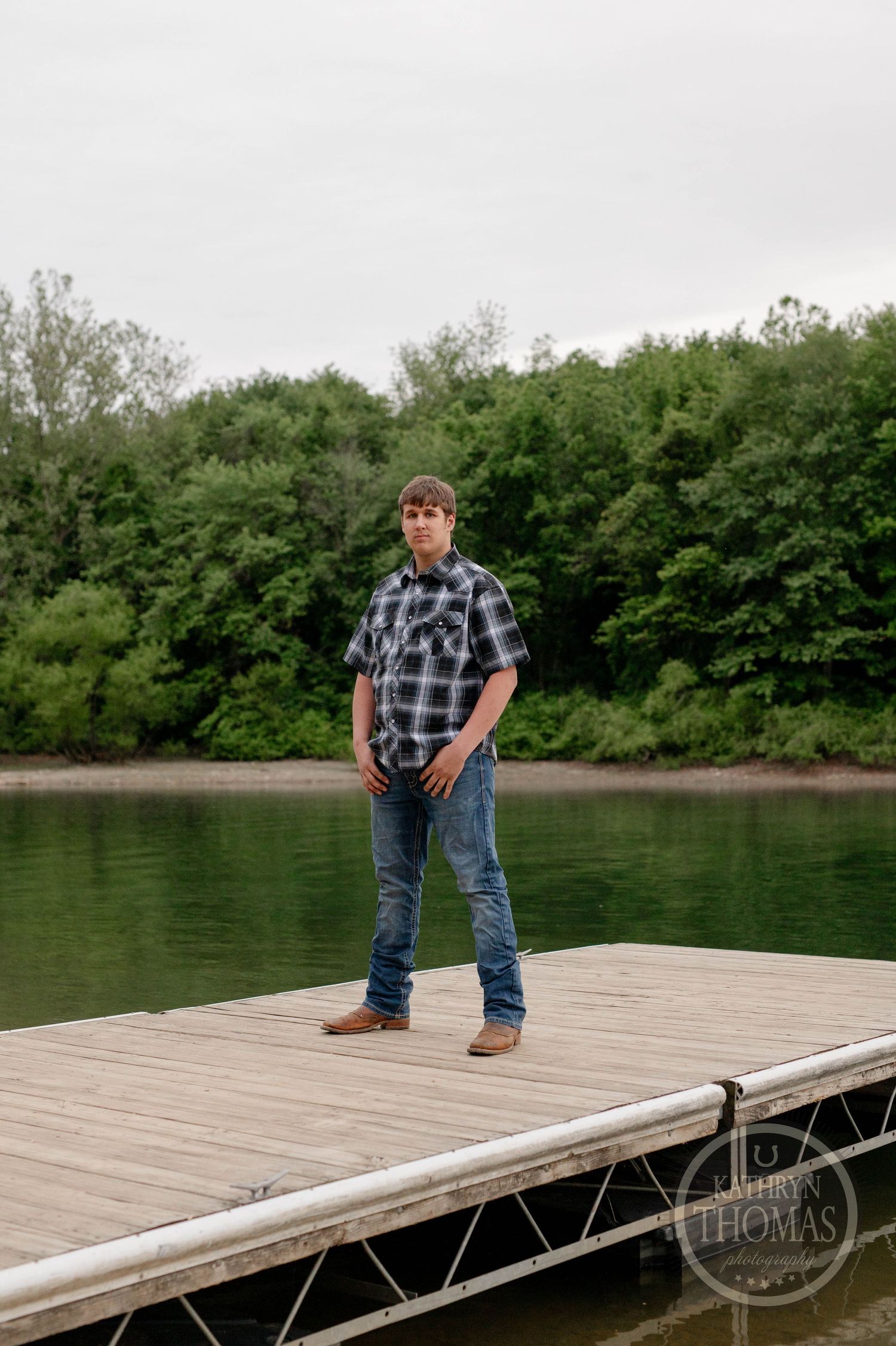 Someone in a plaid shirt and jeans stands on a wooden dock at the edge of a tranquil lake surrounded by trees.