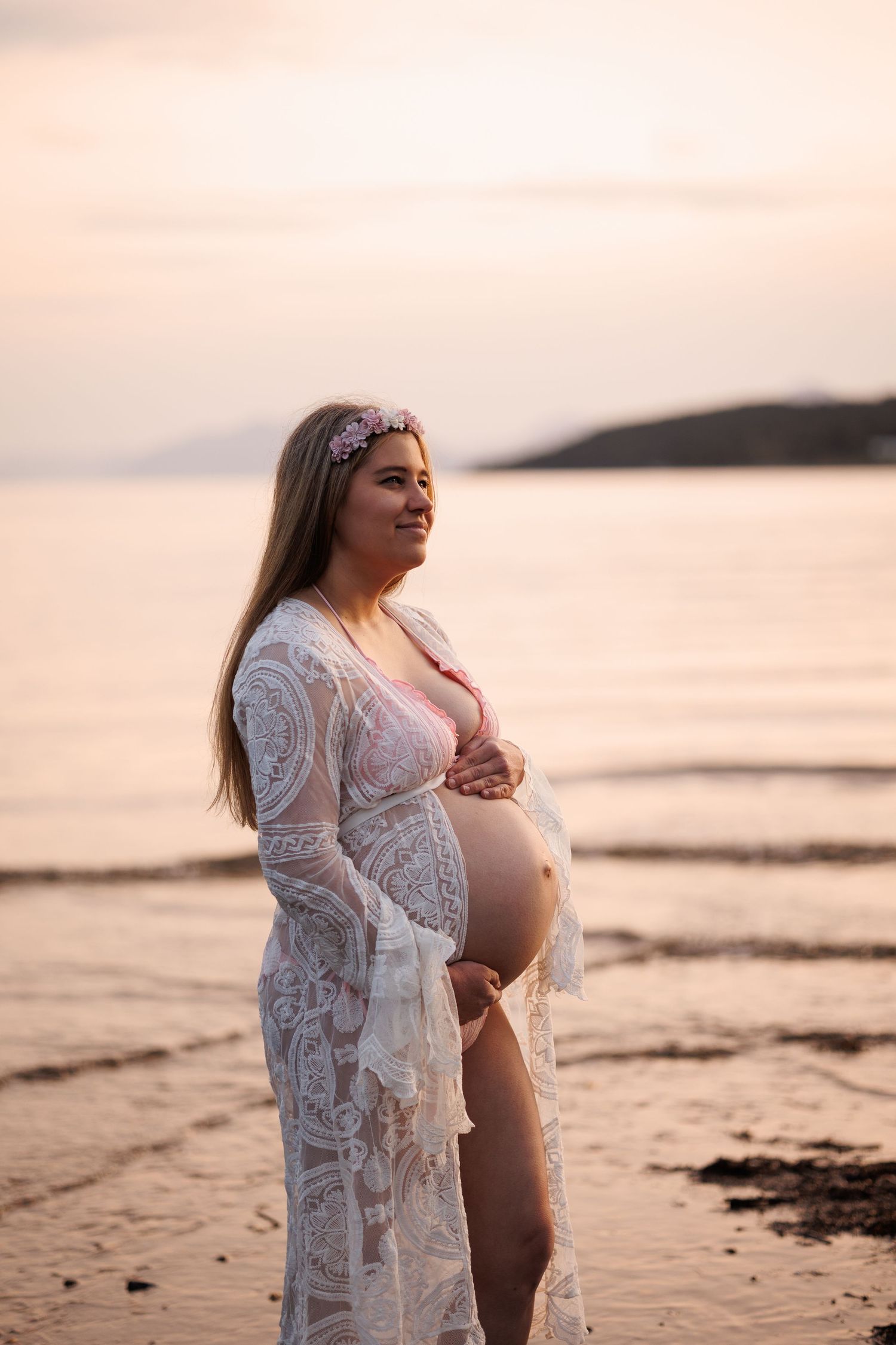 Silhouetted profile of a pregnant belly draped in delicate white lace against golden sunlight.