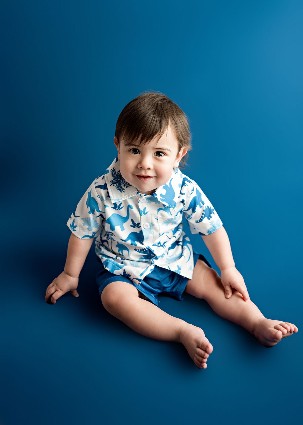 Child in blue and white print shirt and blue shorts sits against solid blue background with bright smile.
