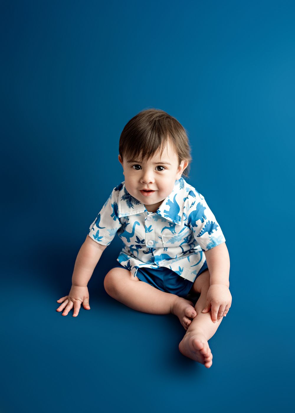 Small child wearing a blue and white patterned shirt sits on a solid blue background with a joyful expression.