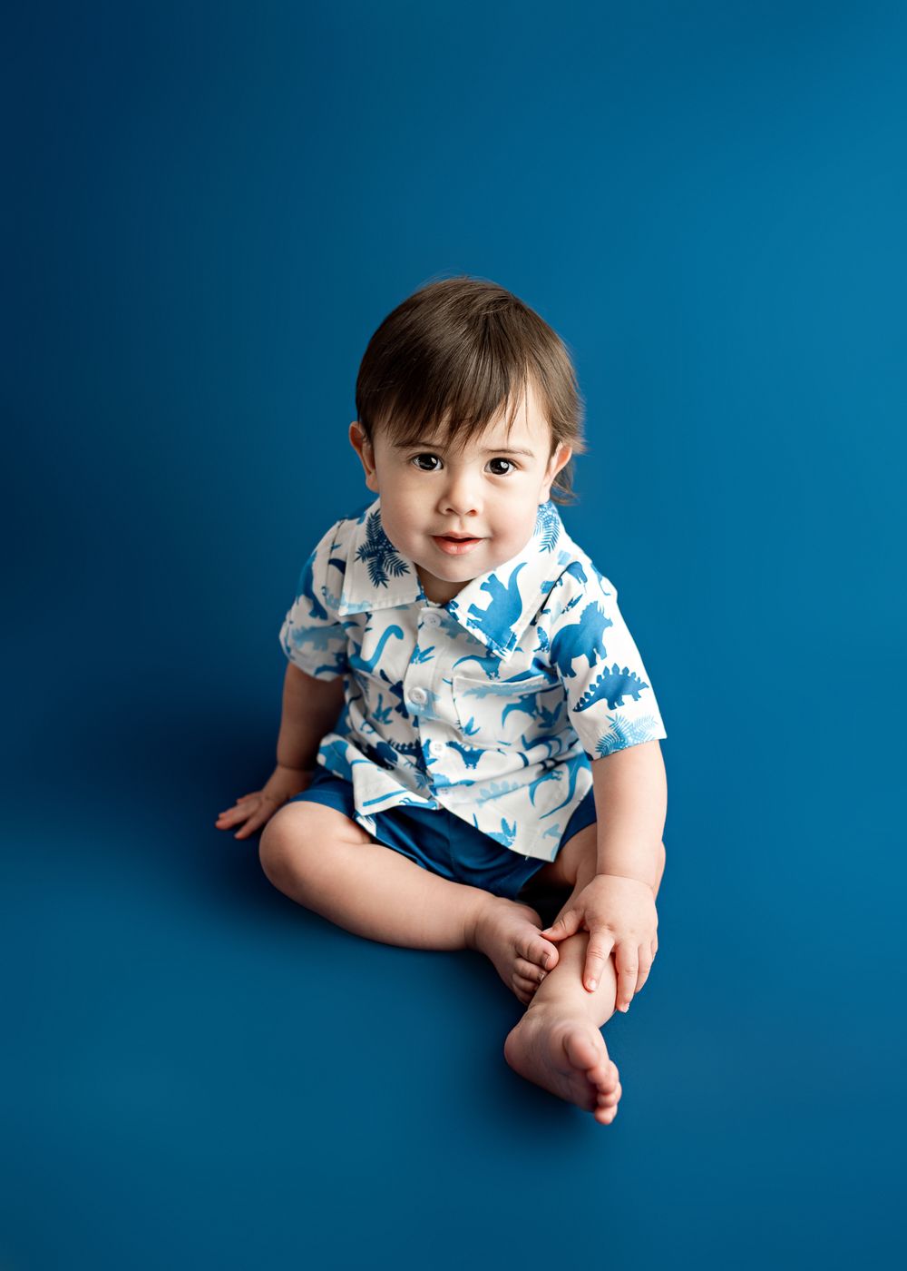 Small child wearing blue and white shark print shirt sits on solid blue background during studio photo session.
