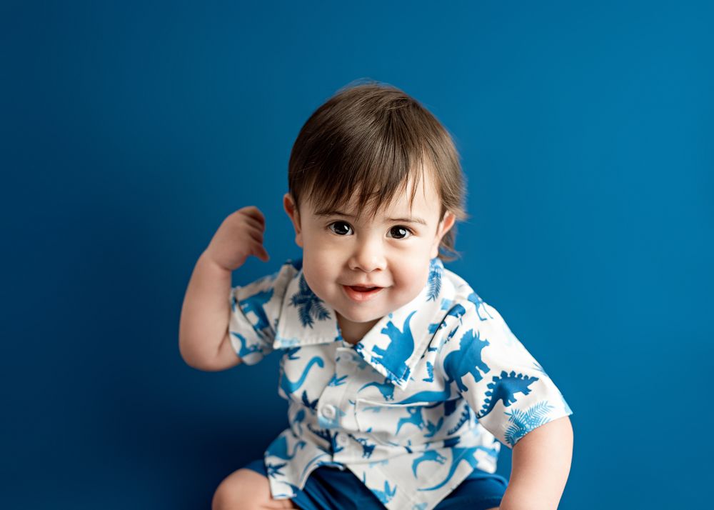 Happy baby wearing white and blue patterned shirt sitting against bright blue background.