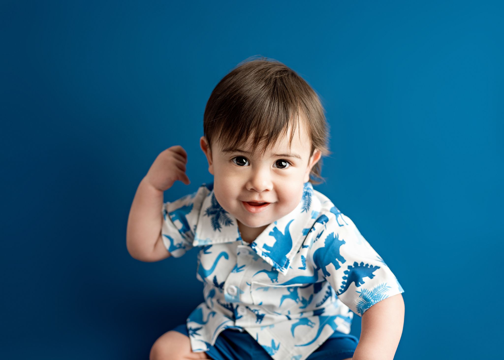 Happy baby wearing white and blue patterned shirt sitting against bright blue background.
