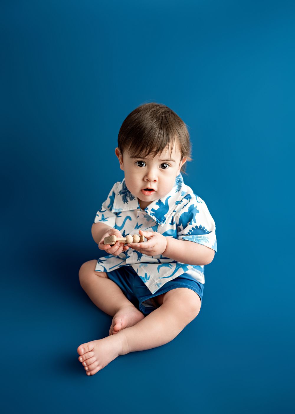 A young child in a patterned blue and white shirt and navy shorts sits on a bright blue background.