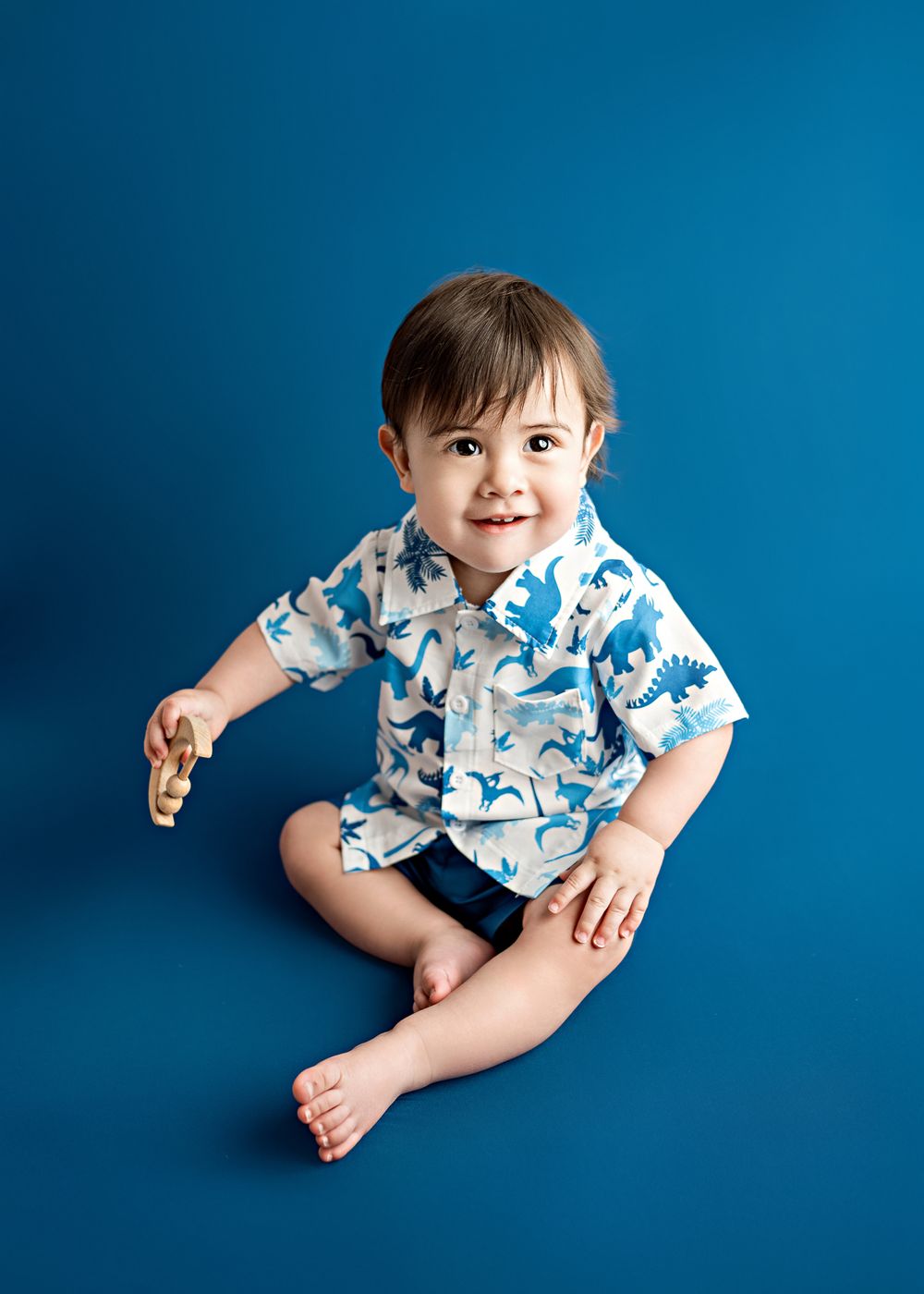 Toddler wearing blue and white patterned shirt sitting against blue backdrop during studio portrait session.
