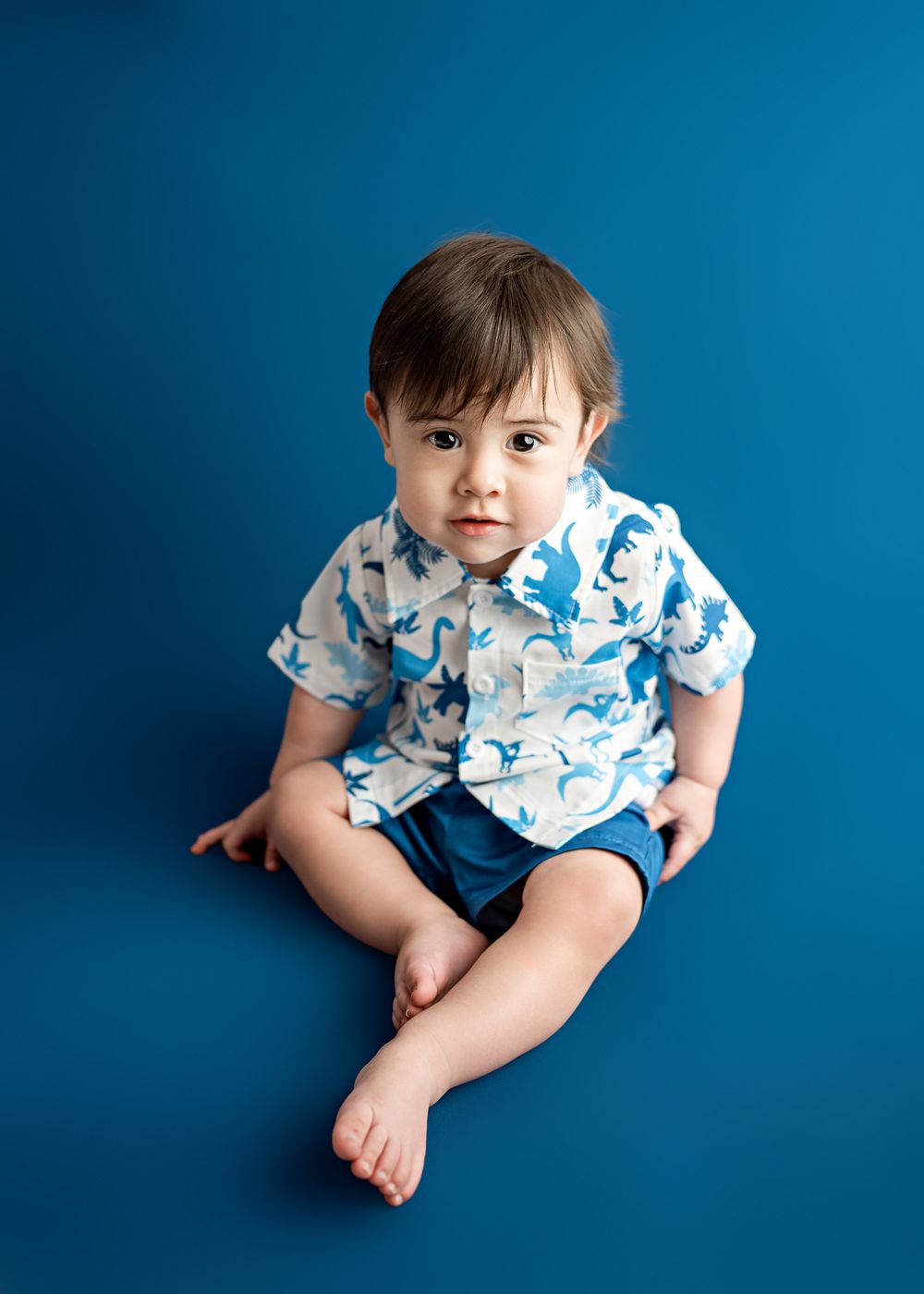 A young child wearing a white and blue patterned shirt sits on a blue background with bare feet.