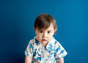 Young child wearing a white and blue patterned shirt stands against a bright blue background.