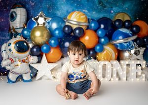 Space-themed first birthday party setup with balloon arch featuring astronauts, planets, and white 'ONE' lettering decoration.