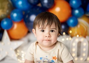 Toddler sits in front of blue and orange balloon decorations and star-shaped lights at a birthday celebration.