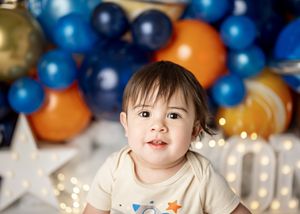 Small child wearing white shirt with stars sitting in front of festive blue and orange balloon decorations.