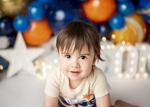 A toddler in a light colored shirt sits in front of festive blue and orange balloon decorations.