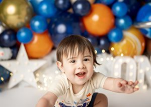 Young child smiling amid festive party decorations including blue, gold and orange balloons with twinkling lights.
