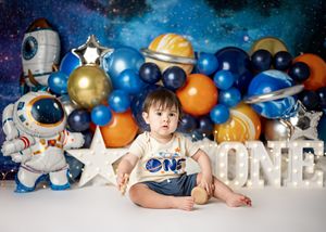 Space-themed first birthday setup with astronaut decorations, colorful balloons, silver stars, and 'ONE' letters against starry backdrop.