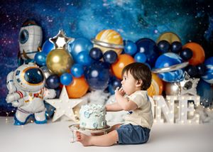 Toddler sits against space-themed backdrop with balloons, stars, and astronaut decorations for first birthday celebration.