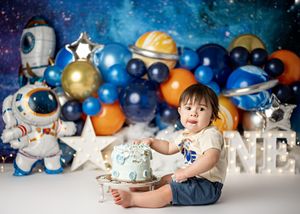 Space themed first birthday party setup with astronaut decorations, balloons, and a blue and silver cake against starry backdrop.
