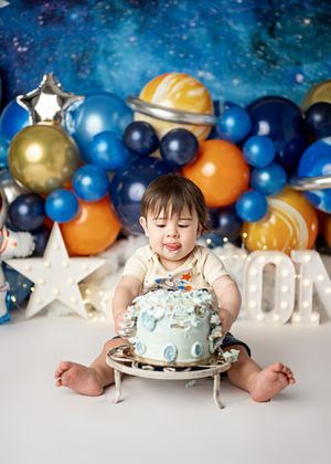 A cake smash photo with blue and orange balloons, light-up stars and a decorated cake set against a cosmic backdrop.