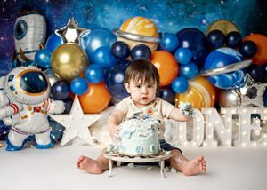 A baby in a tutu sits against a backdrop of blue and gold balloons, silver stars, and astronaut decorations for space theme party.