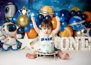 A baby sitting among space-themed decorations with blue and gold balloons, stars, and a ONE sign for their first birthday party.