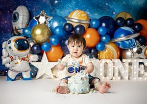 Space-themed first birthday cake smash setup with balloons, stars and astronaut decorations against a cosmic backdrop.